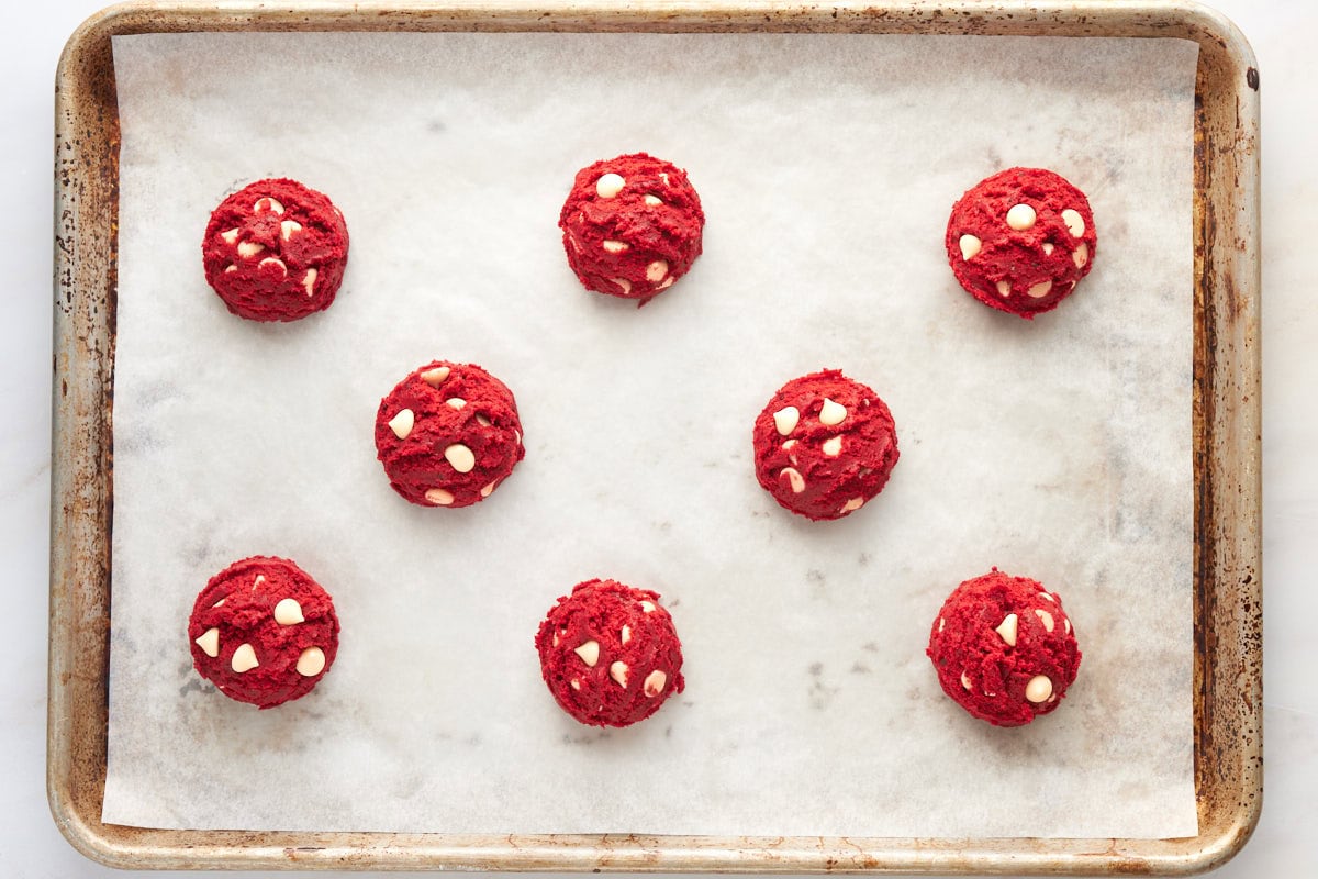 Scooped portions of red velvet cookie dough with white chocolate chips arranged on parchment lined baking tray.